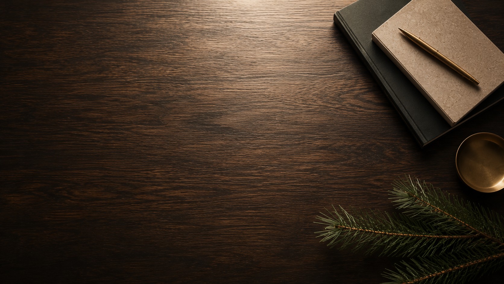 Notebooks and a brass pen on a dark wooden desk with pine branches.